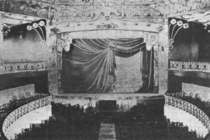 Schwarzes und weißes Foto des Auditoriums des Theatre Royal in London, das eine Bühne mit einem Vorhang, Geländern und ornamentalen Wanddesigns zeigt, mit einigen sitzenden Zuschauern und Text am unteren Rand.