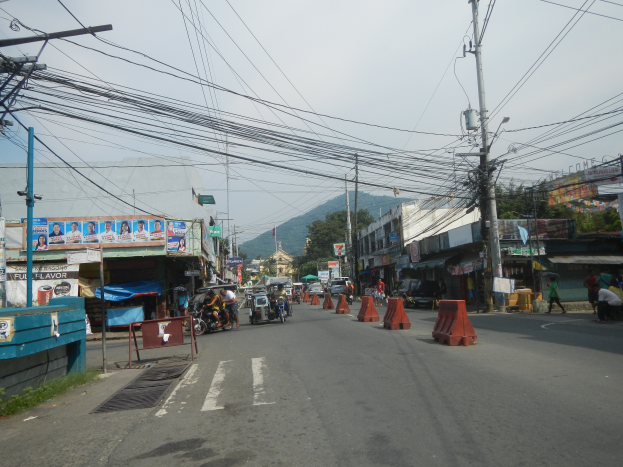 Eine Stadtstraße mit Strommasten, Fahrzeugen, Fußgängern, Gebäuden, Werbetafeln, Bäumen, Hügeln und einem klaren blauen Himmel.
