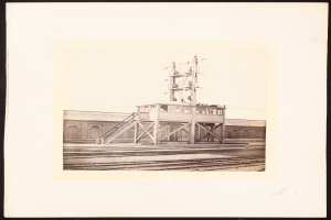 Ein Schwarz-Weiß-Foto eines Bahnhofs mit einer zentralen Signalbox, umgeben von Bahnschienen und einem Gebäude im Hintergrund, mit Text unten.