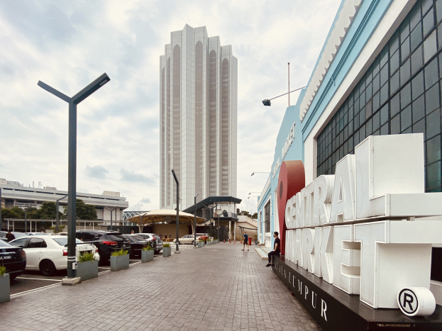 Ein belebter zentraler Markt in Kuala Lumpur, Malaysia, mit hohen Gebäuden, Straßenlaternen, Straßenschildern, Kraftfahrzeugen, Fußgängern, Namensschildern, Bäumen und einem bewölkten Himmel.