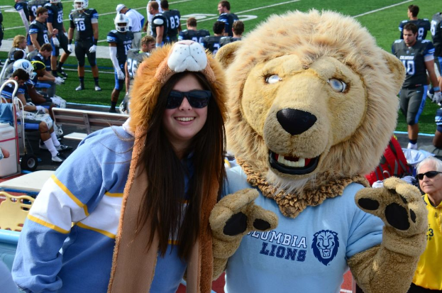 Eine Frau steht neben einer Person in einem Löwenkostüm, mit Spielern auf dem Boden dahinter, einigen Menschen auf einer Bank links und Stühlen neben der Bank.