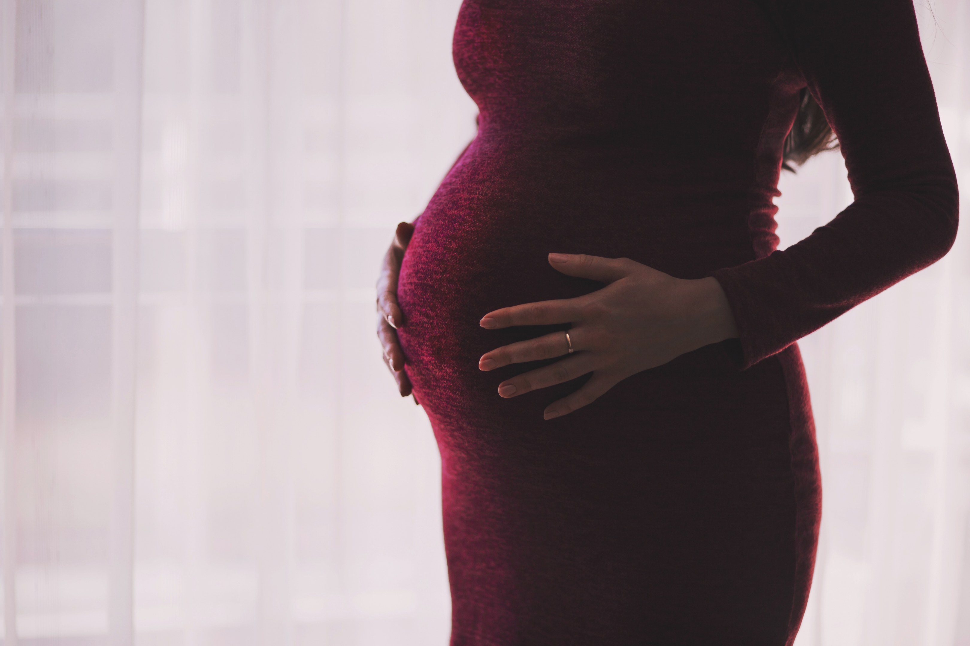 Schwangere Frau in einem roten Kleid, die vor einem Fenster steht und ihren Bauch berührt, mit einem leicht unscharfen Hintergrund.