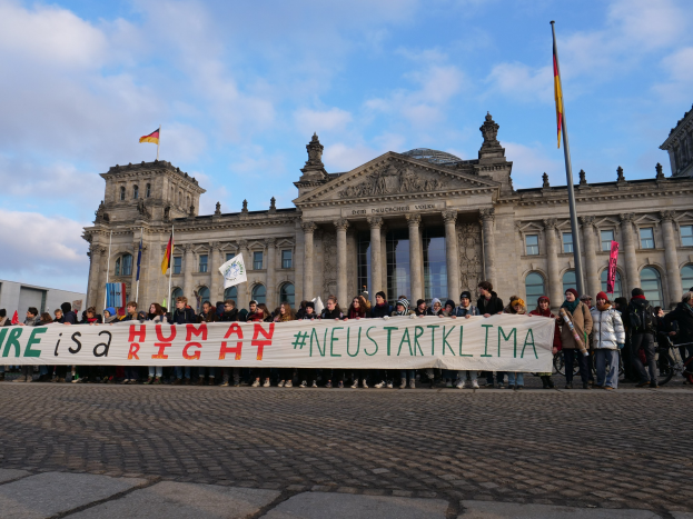 Eine Gruppe von Menschen steht vor dem Reichstagsgebäude in Berlin und hält eine Fahne mit der Aufschrift "Wir sind ein Menschenrecht" in den Händen, wobei Details der Architektur des Gebäudes und Flaggen im Hintergrund zu sehen sind.