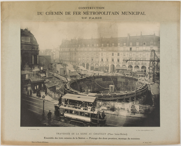Schwarzes und weißes Foto einer Pariser Stadtstraße im Bau mit einem Zug auf den Schienen, umgeben von Gebäuden, Polen und Menschen; Text oben lautet "Construction du Chemin de Fer Metropolitain Municipal de Paris".