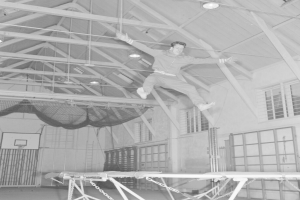 Ein Mann in der Luft springt auf einem an der Decke hängenden Trampolin in einer Turnhalle, mit ausgestreckten Armen und gebeugten Knien, dargestellt in Schwarz-Weiß.