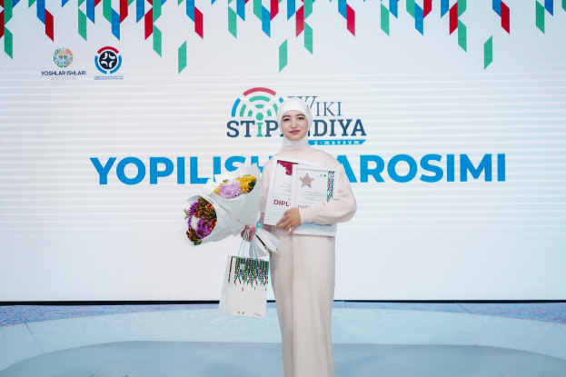 Eine Frau steht auf einer Bühne, hält in einer Hand einen Blumenstrauß und in der anderen ein Zertifikat, während hinter ihr ein Banner mit der Aufschrift 'Yopilisi Sarosimi' hängt, was darauf hinweist, dass sie die Gewinnerin des Yopilisi Sarosimi Awards ist.