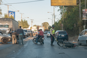 Eine Gruppe von Menschen um ein verunglücktes Motorrad auf der Straße herumstehend mit mehreren Fahrzeugen, darunter ein Lastwagen, und einem Hintergrund aus Bäumen, Polen, Lampen, Brettern und Himmel.